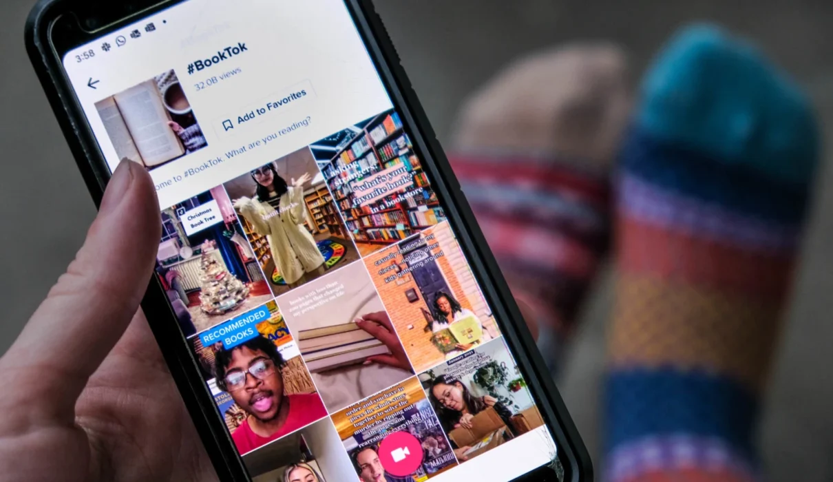 smartphone affichant l'application TikTok, ouvert sur une vidéo d’une jeune femme en train de montrer un livre, avec une pile de romans colorés en arrière-plan, ambiance chaleureuse et lumineuse