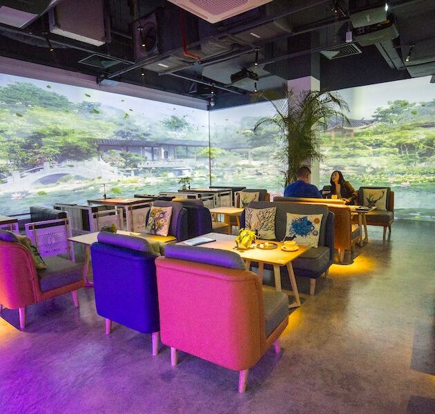 Une photo d’ambiance d’un café tamisé avec des projections artistiques immersives sur les murs, des clients interagissant avec des œuvres digitales ou scannant un mur en réalité augmentée, le tout dans une atmosphère chaleureuse et créative.