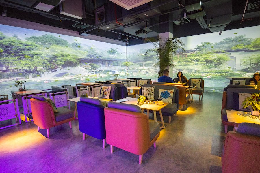 Une photo d’ambiance d’un café tamisé avec des projections artistiques immersives sur les murs, des clients interagissant avec des œuvres digitales ou scannant un mur en réalité augmentée, le tout dans une atmosphère chaleureuse et créative.
