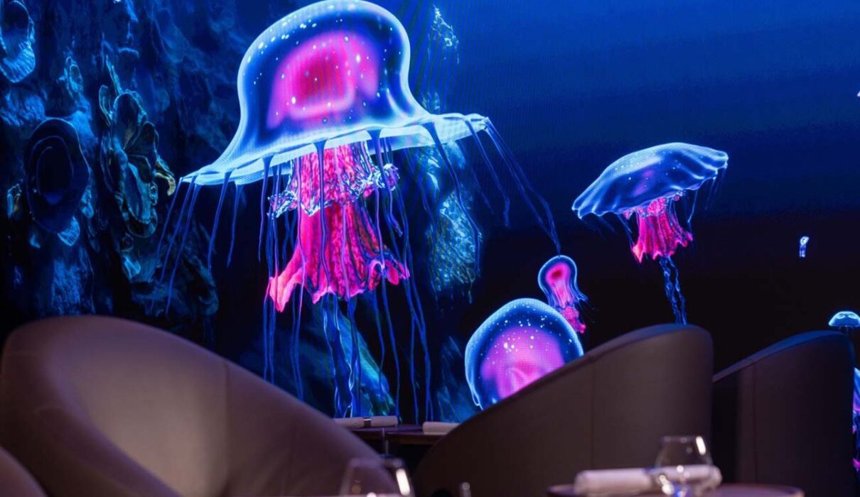 Une grande table élégante plongée dans une lumière tamisée, avec projection immersive sur les murs (océan ou forêt), un plat artistique au centre et des convives émerveillés