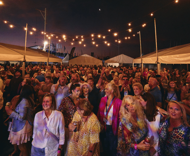 foule de festivaliers stylés sous un ciel coloré, avec des tenues audacieuses et des stands de marques, capturant l’énergie, la créativité et le côté commercial du street style